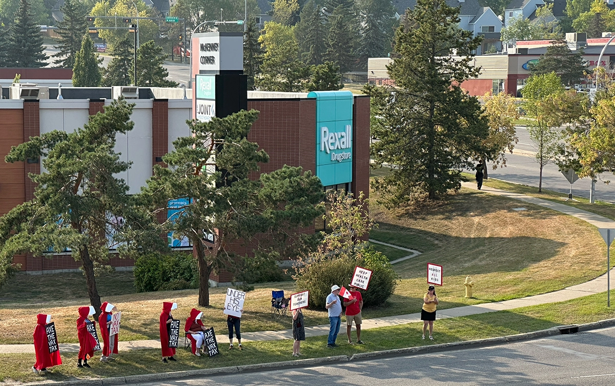 Wide view of a small protest in St. Albert with people standing along a roadside holding signs, including several wearing red Handmaid’s Tale–style outfits.