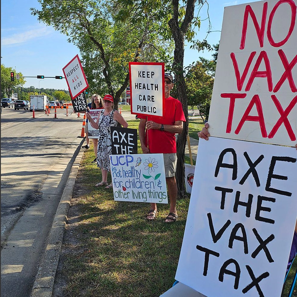 Protesters holding signs reading “Axe the Vax Tax” and “Keep Health Care Public,” viewed from the roadside with a large sign in the foreground.