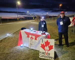 Volunteers staffing a Forever Canadian petition table at dusk, with flags and materials set up along a roadside.