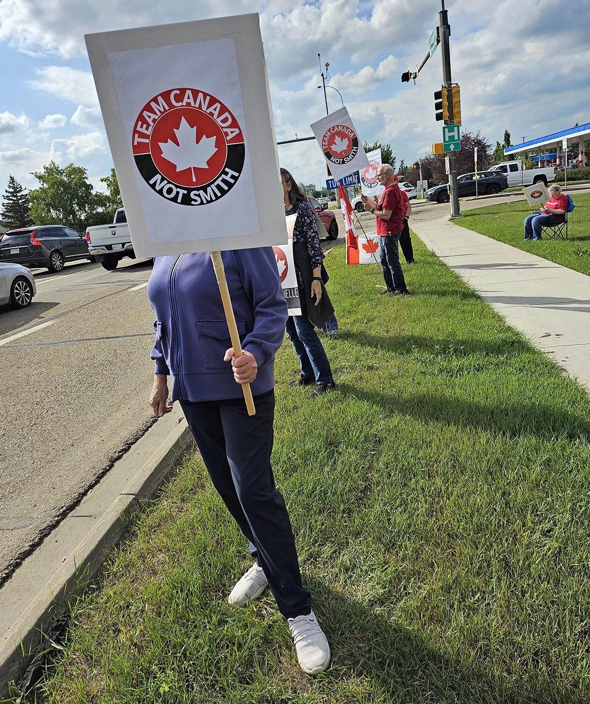Members of our group stand along a sidewalk in St. Albert holding signs and a Canadian flag during a roadside rally.