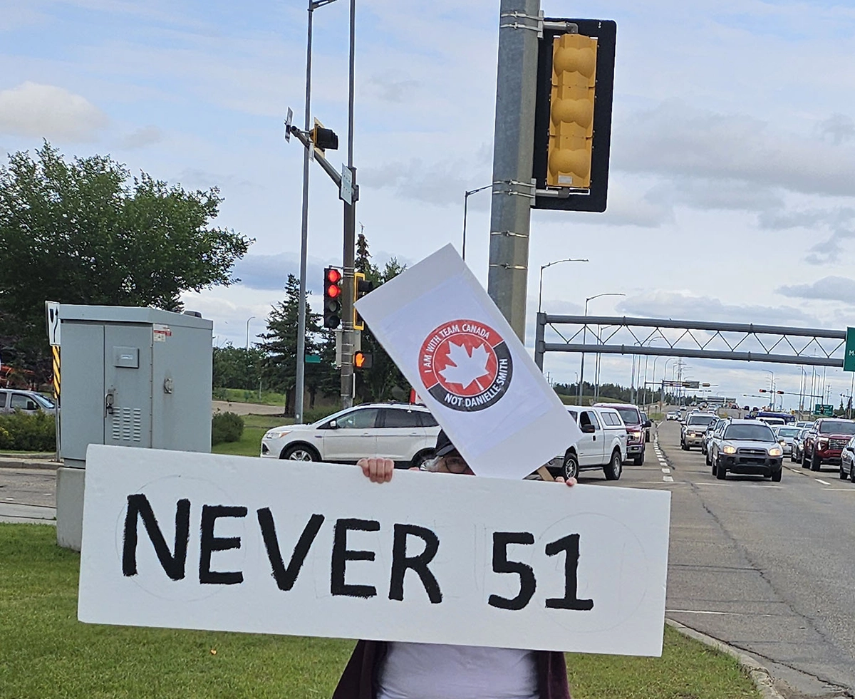 A participant holds a “Never 51” sign during a roadside rally with traffic in the background.