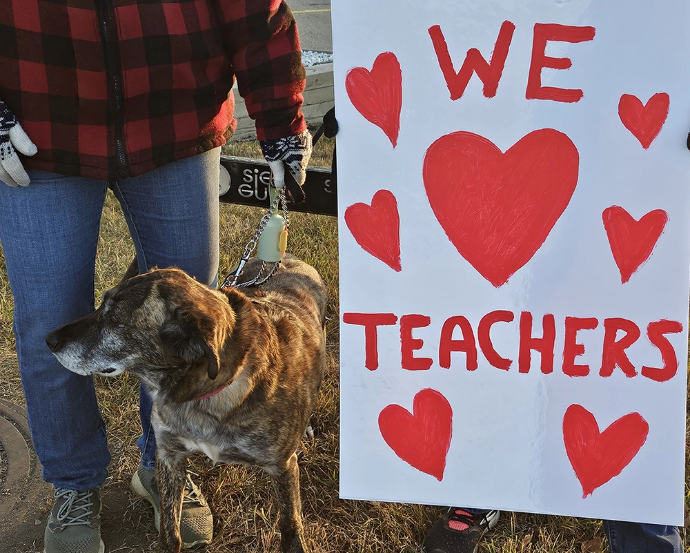 Person with dog standing beside sign reading “We love teachers” at roadside rally