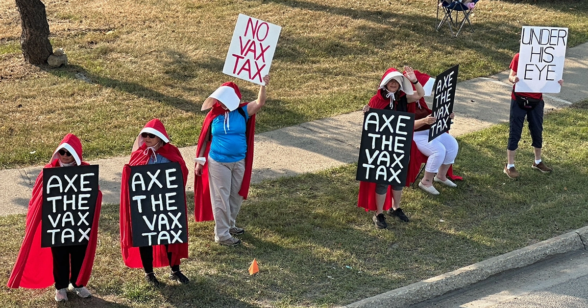 Close view of protesters, several in Handmaid’s Tale–style outfits, holding “Axe the Vax Tax” signs along a sidewalk.